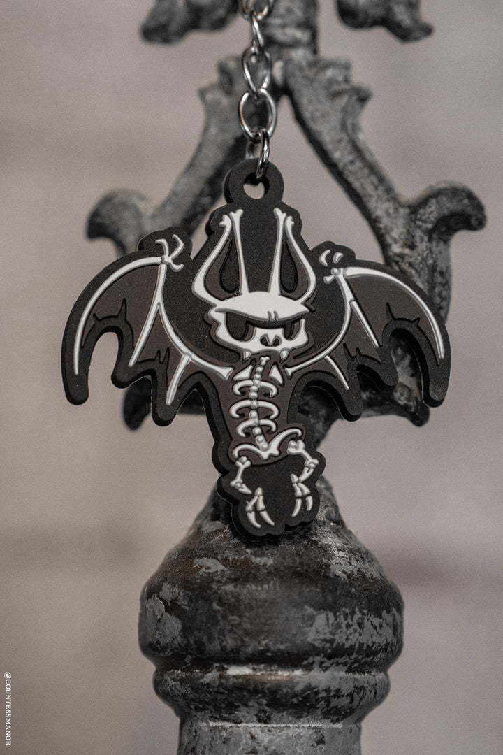 Bat-shaped keychain with skeleton design on a metallic stand against a blurred background