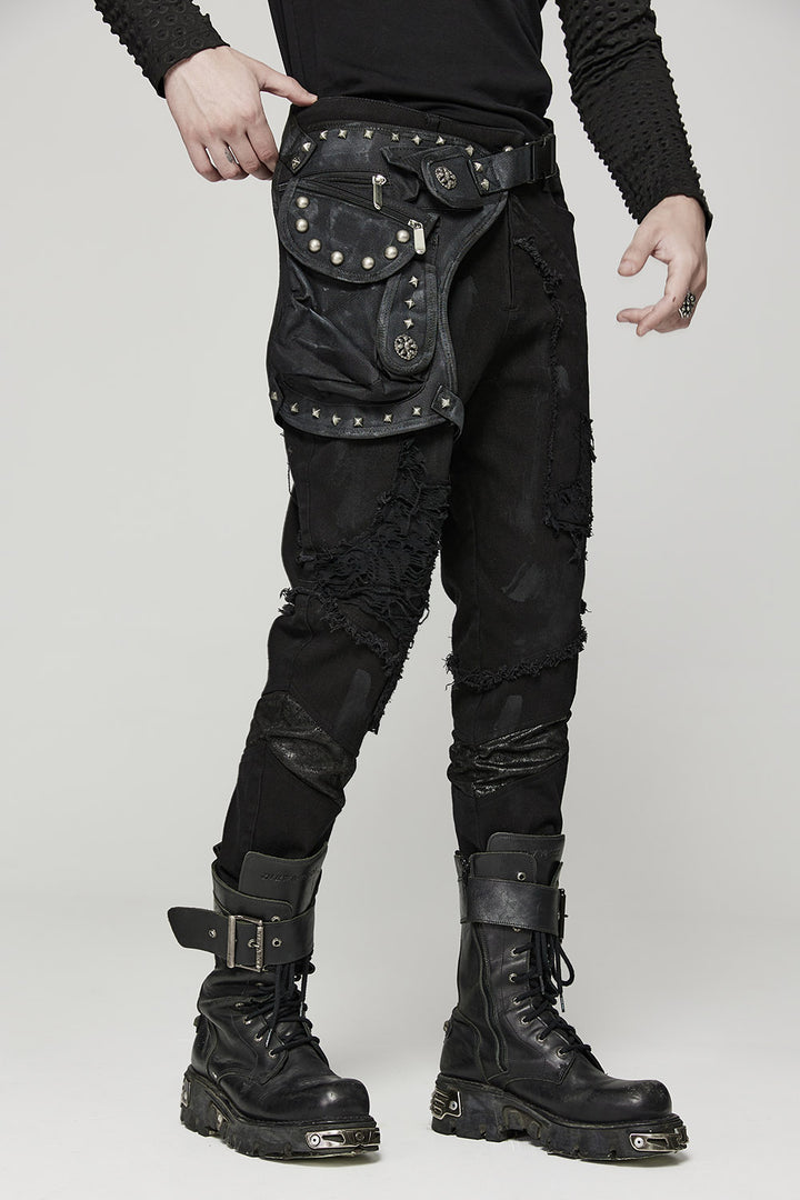 A person wearing a black leg harness bag with distressed pyramid studs and metal skull hardware, featuring zippered pockets, leg and waist straps. The item is hand-painted, and the person is dressed in all black with black boots.