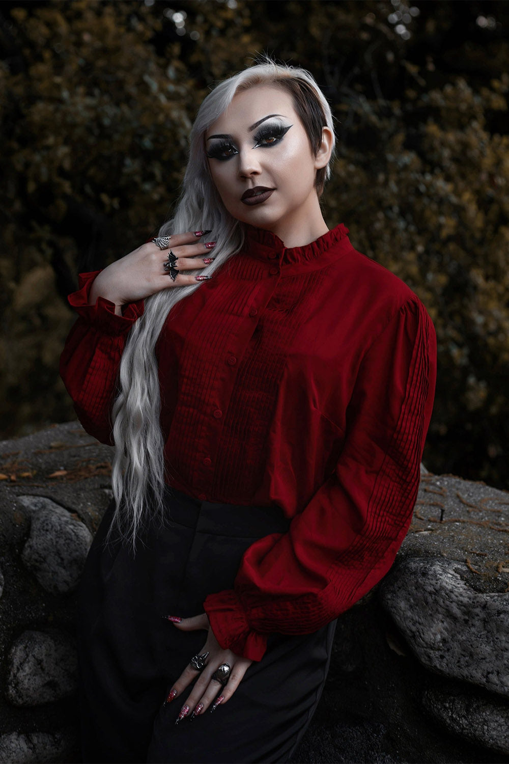 Person wearing a red blouse with dark hair and makeup, standing against a blurred natural background.