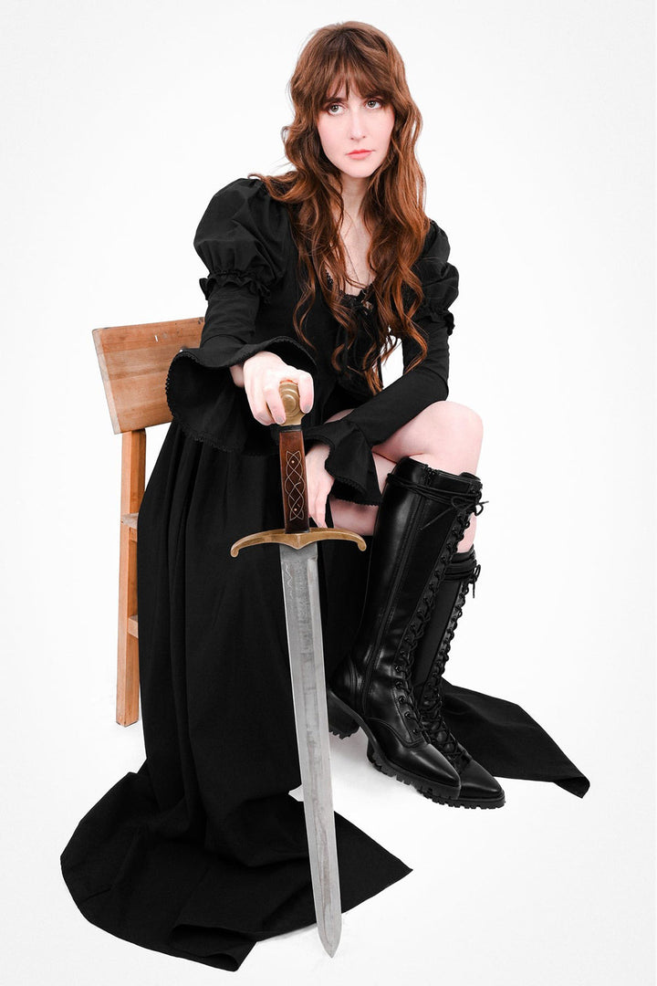 Woman in a black duster dress holding a sword, sitting on a chair against a white background