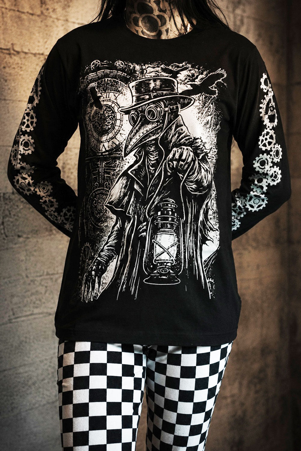 Person wearing a black long-sleeve shirt with a detailed graphic design of a steampunk character holding a lantern, standing against a textured wall.