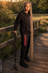 A person standing on a wooden boardwalk outdoors at sunset, wearing a black hoodie and black sweatpants with red Gothic-style lettering that reads 'Langst' down the left leg. The person holds a cane and has long blonde dreadlocks.