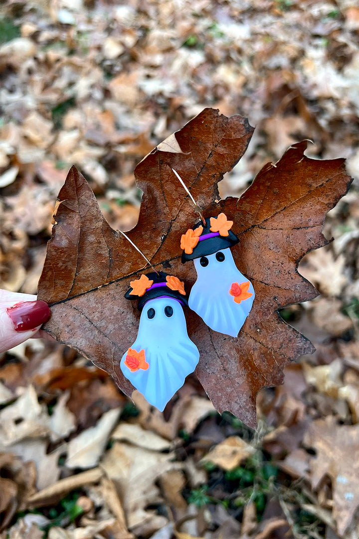 Two ghost-shaped earrings with orange accents on a leaf in a natural setting.