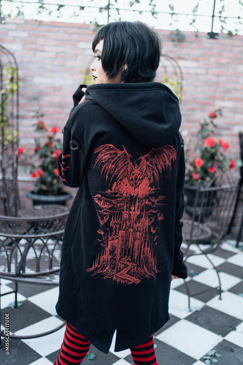 A person wearing a black hooded coat with a large red graphic print on the back, standing on a black-and-white checkered patio with a brick wall and potted red flowers in the background.