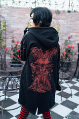 A person wearing a black hooded coat with a large red graphic print on the back, standing on a black-and-white checkered patio with a brick wall and potted red flowers in the background.