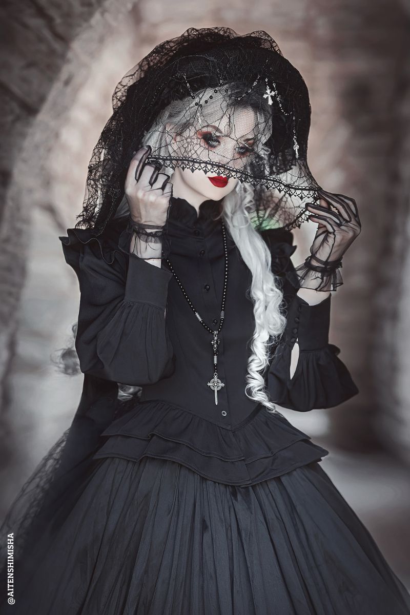 A gothic-style model wearing a black layered ruffle top, a large black lace hat with a veil, and a cross necklace, posed against a dark, atmospheric stone background.