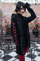 A person wearing a black hooded jacket with red bat motifs on the sleeve, a black top, and red-and-black striped tights, standing in an outdoor patio setting with brick walls, metal chairs, and potted red flowers.