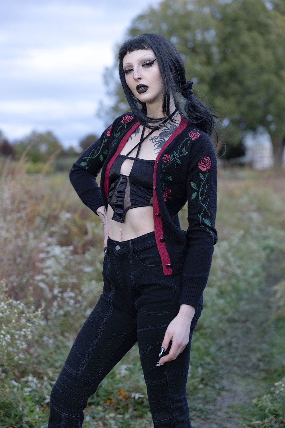 A woman posing outdoors in a gothic style, wearing a black cardigan with red rose embroidery and red trim, a black strappy crop top, and black jeans.