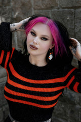Person with pink hair wearing a black and orange striped sweater against a stone wall with ghost earrings