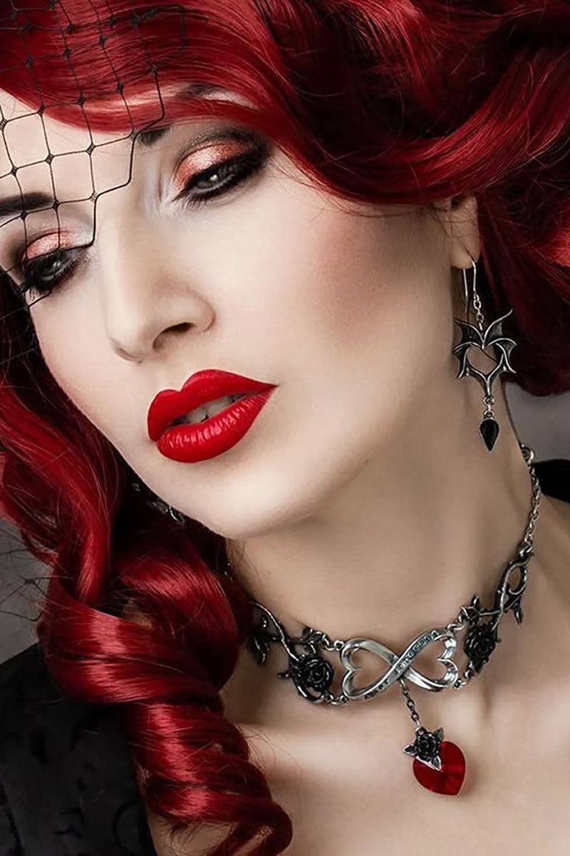 Close-up of a woman with red hair and red lipstick wearing a silver necklace with a heart pendant and batwing earrings