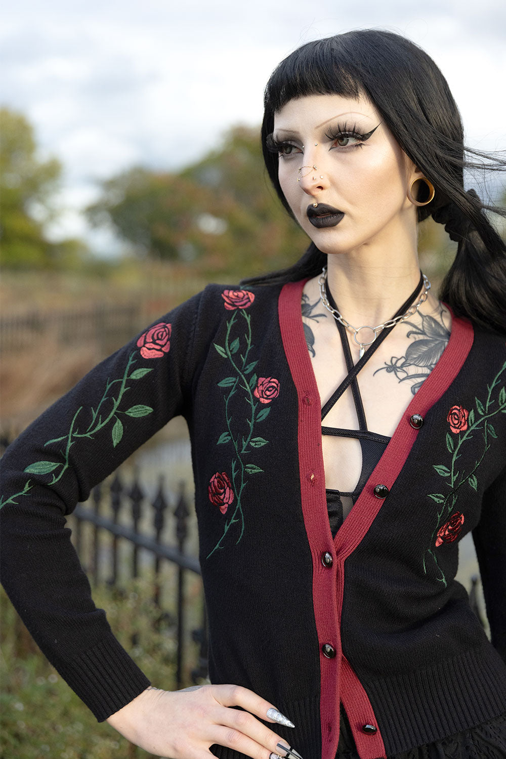 A woman with gothic makeup and a black wig wearing a black cardigan with red trim and green rose embroidery, standing outdoors in front of a blurred fence and trees.