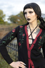A woman with gothic makeup and a black wig wearing a black cardigan with red trim and green rose embroidery, standing outdoors in front of a blurred fence and trees.