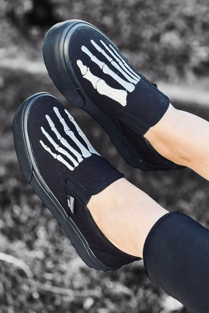 Black slip-on shoes with white skeleton design on a blurred outdoor background
