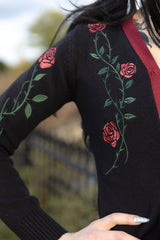 A close-up of a person wearing a black knit sweater with embroidered red roses and green vines, paired with a dark red ribbed V-neck trim, set against a blurred outdoor background.