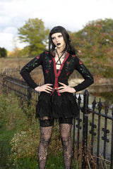 A gothic-style model stands outdoors next to a black metal fence, wearing a black long-sleeve cardigan with red trim and rose embroidery, a matching black ruffled mini skirt, black lace tights, and a black wig.