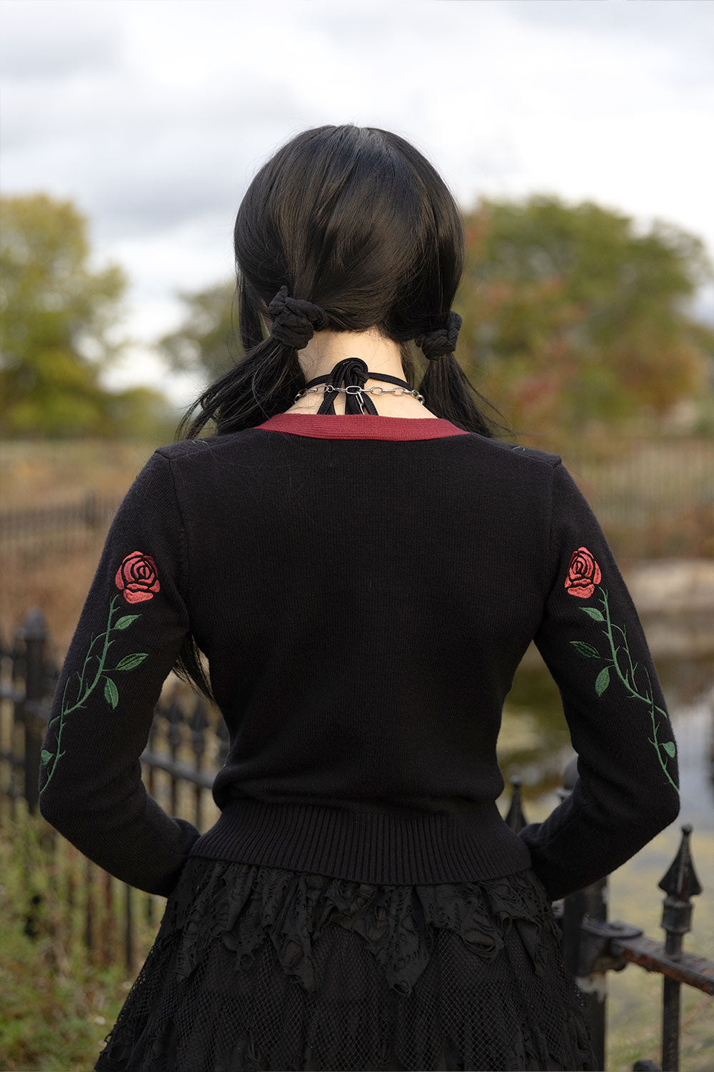 A person viewed from behind wearing a black long-sleeve sweater with embroidered red roses and green leaves on the sleeves, a black lace skirt, and a black choker necklace. The background shows an outdoor scene with trees and a fence.
