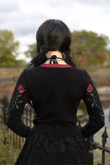 A person viewed from behind wearing a black long-sleeve sweater with embroidered red roses and green leaves on the sleeves, a black lace skirt, and a black choker necklace. The background shows an outdoor scene with trees and a fence.
