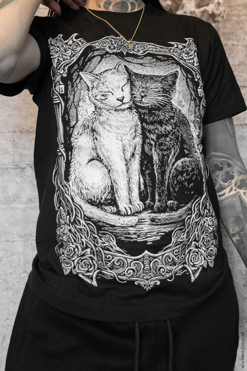 Black t-shirt with a graphic of two cats in a decorative frame worn by a person.