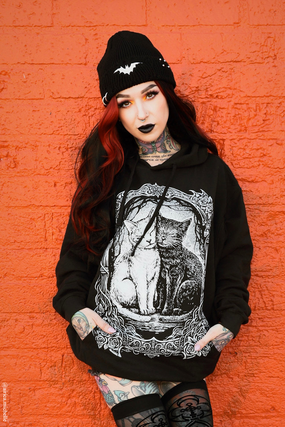 A person with red hair, tattoos, and black lipstick stands against a bright orange brick wall wearing a black beanie with a bat graphic, a black hoodie with a large white cat illustration on the front, and black patterned tights.