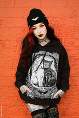 A person with red hair, tattoos, and black lipstick stands against a bright orange brick wall wearing a black beanie with a bat graphic, a black hoodie with a large white cat illustration on the front, and black patterned tights.