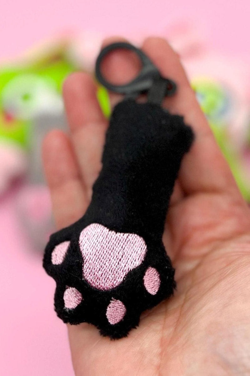 Black cat paw-shaped keychain with pink details held in a hand against a blurred background