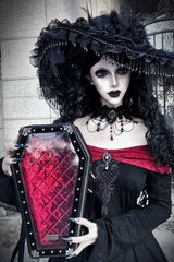 A gothic-style model wearing a large black feathered hat, black lace choker, and a black dress with a red off-the-shoulder strap, holding a coffin-shaped black purse with a red quilted interior.