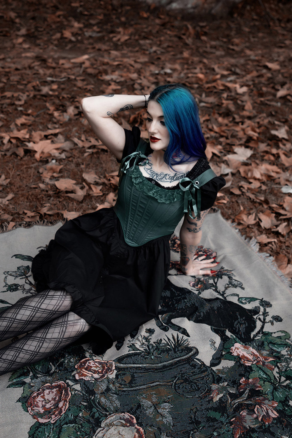 Woman with blue hair sitting on a rug in a forest setting wearing a lace ruffle corset