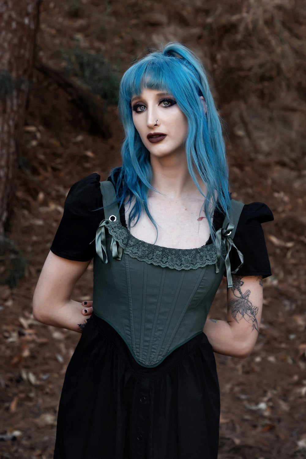 Person with blue hair and green corset top standing in a forest setting