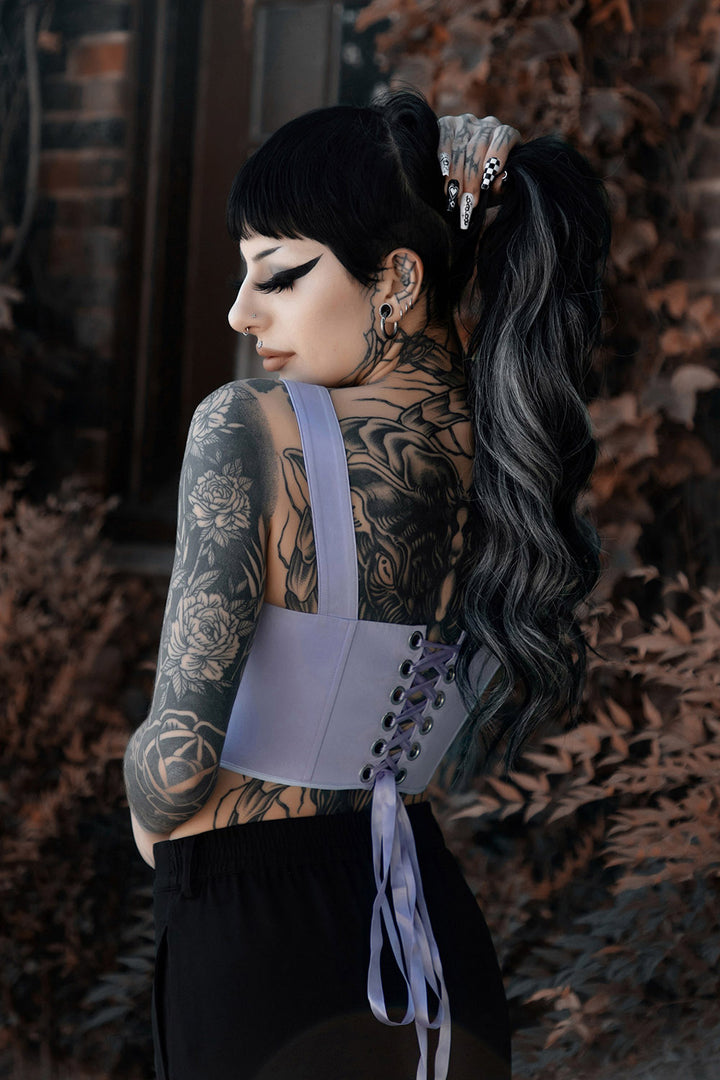 Person wearing a light purple corset with intricate designs, standing against a textured wall.