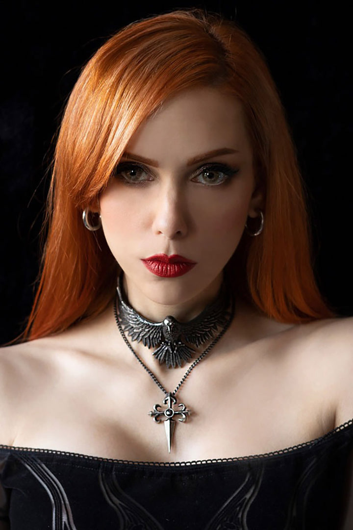 Woman with red hair and red lipstick wearing a black off-shoulder top and a detailed raven choker necklace against a dark background.