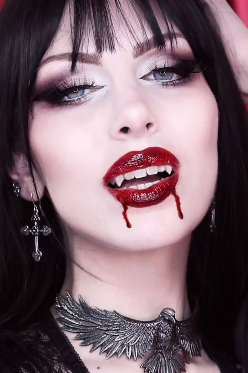 Woman with dark makeup, red lipstick, and a necklace with a bird design.