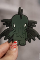 Green Cthulhu magnet held by a hand with red and white nail polish on a gray background