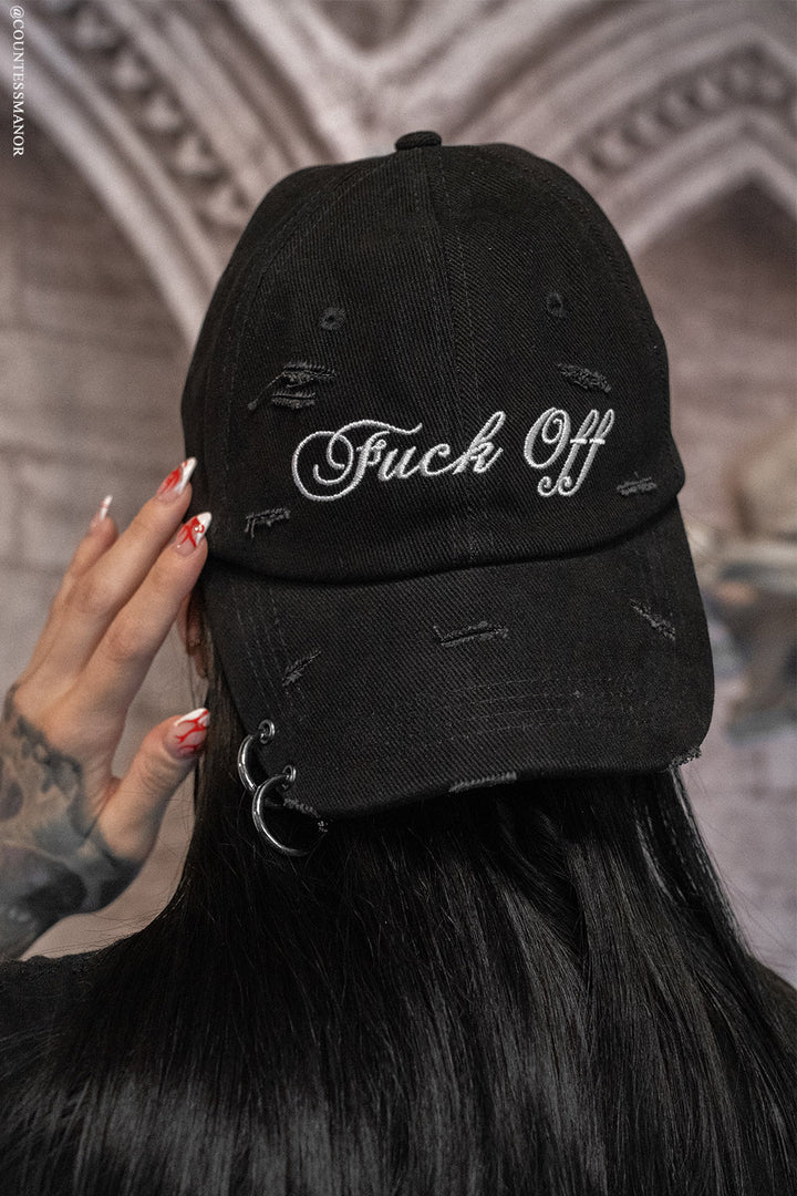 Black cap with 'Fuck Off' text held by a person with tattoos against a blurred background