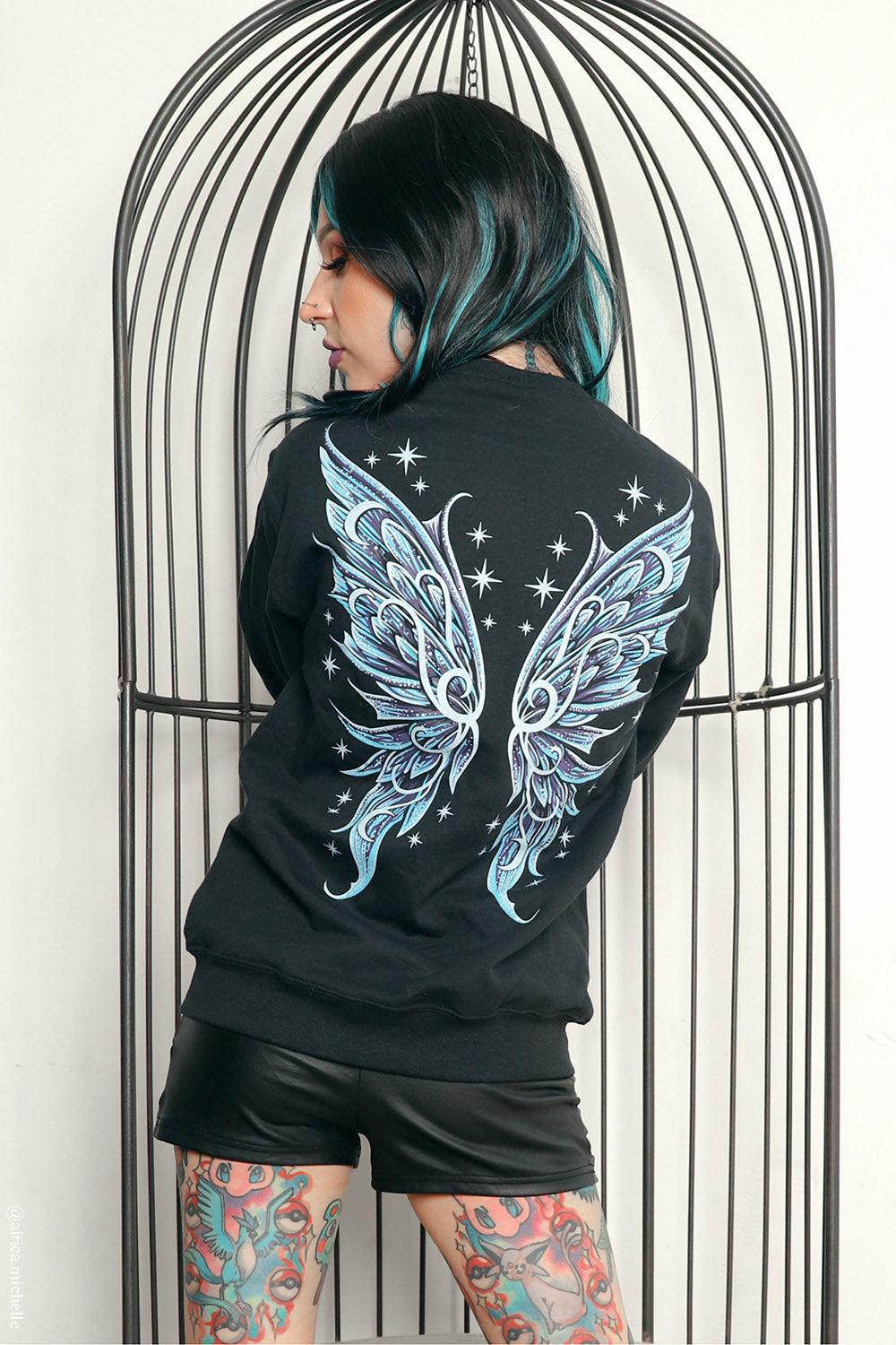 A person with black and teal hair stands in front of a black metal birdcage, wearing a black sweatshirt with a large light blue and white wing graphic on the back, paired with black shorts.