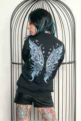 A person with black and teal hair stands in front of a black metal birdcage, wearing a black sweatshirt with a large light blue and white wing graphic on the back, paired with black shorts.