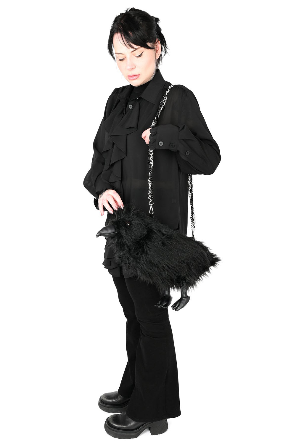 A model wearing a black gothic outfit, including a long-sleeve button-up shirt, flared pants, and black boots, carrying a black raven-shaped shoulder bag with a silver chain strap against a solid white background.