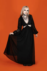 A model with platinum blonde hair and gothic makeup wearing a long black dress with sheer long sleeves, cutouts at the waist, and a sheer black overlay, posing against a solid orange background.