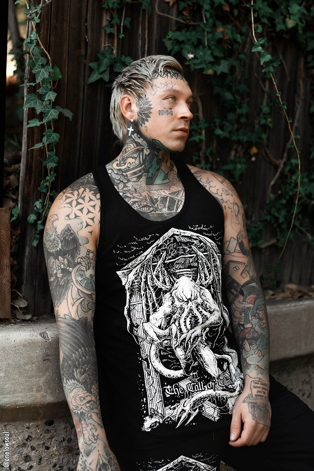 Person with tattoos wearing a black tank top with a graphic design, sitting outdoors.