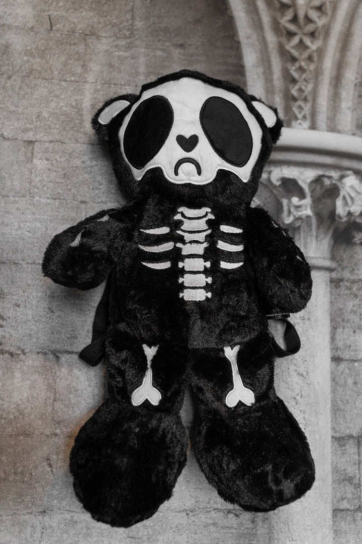 Black skeleton plush teddy bear backpack toy against a stone wall background