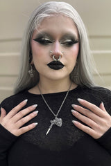 Person with silver hair and dark makeup wearing a black top and gothic jewelry 