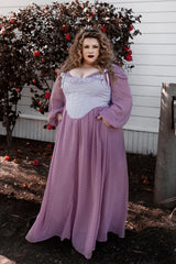Woman in a purple dress and corset standing in front of a white building with a bush bearing red fruits.