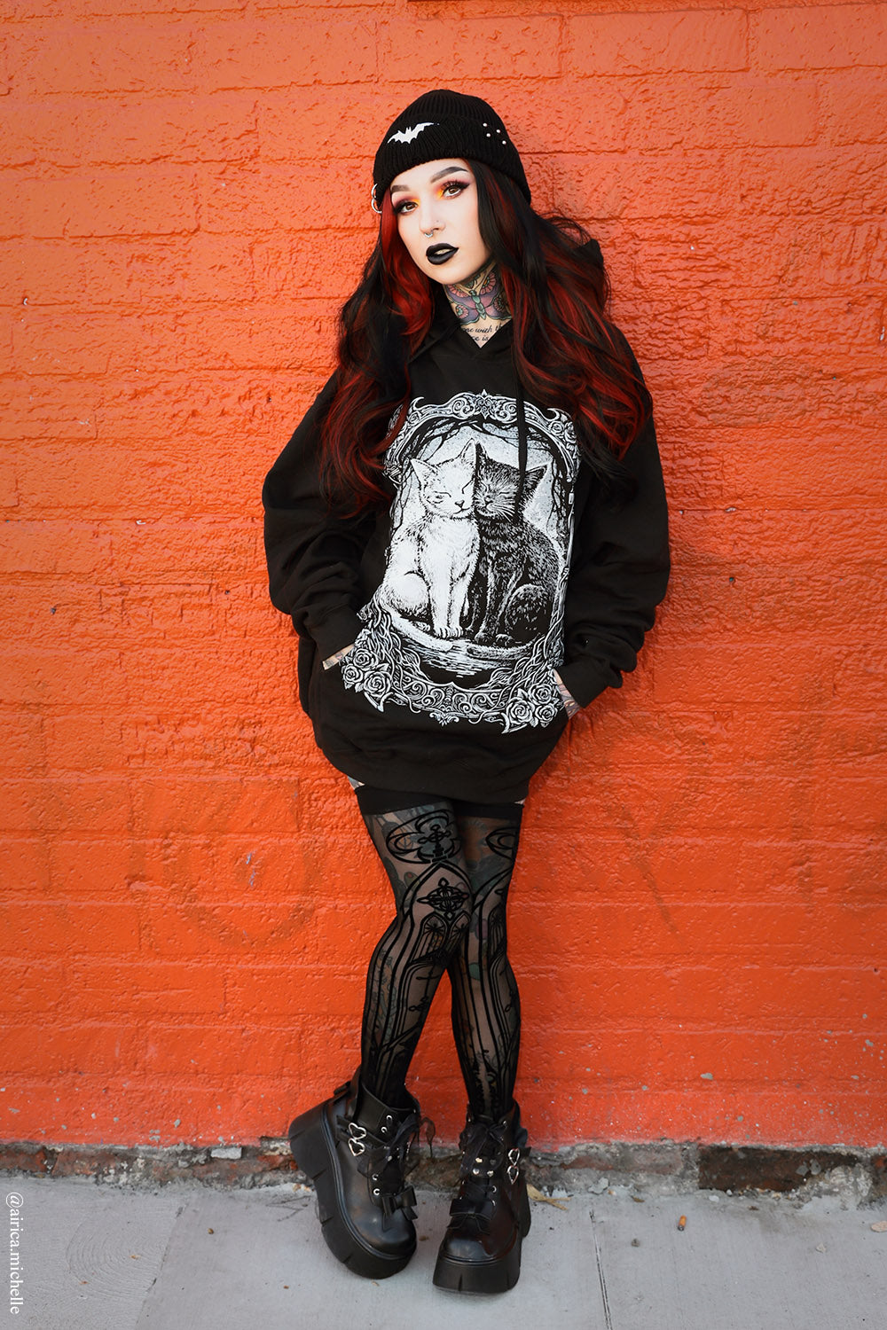 A goth-style model with long red hair and black makeup stands against a bright orange brick wall, wearing a black graphic hoodie, black tights, black boots, and a black beanie.
