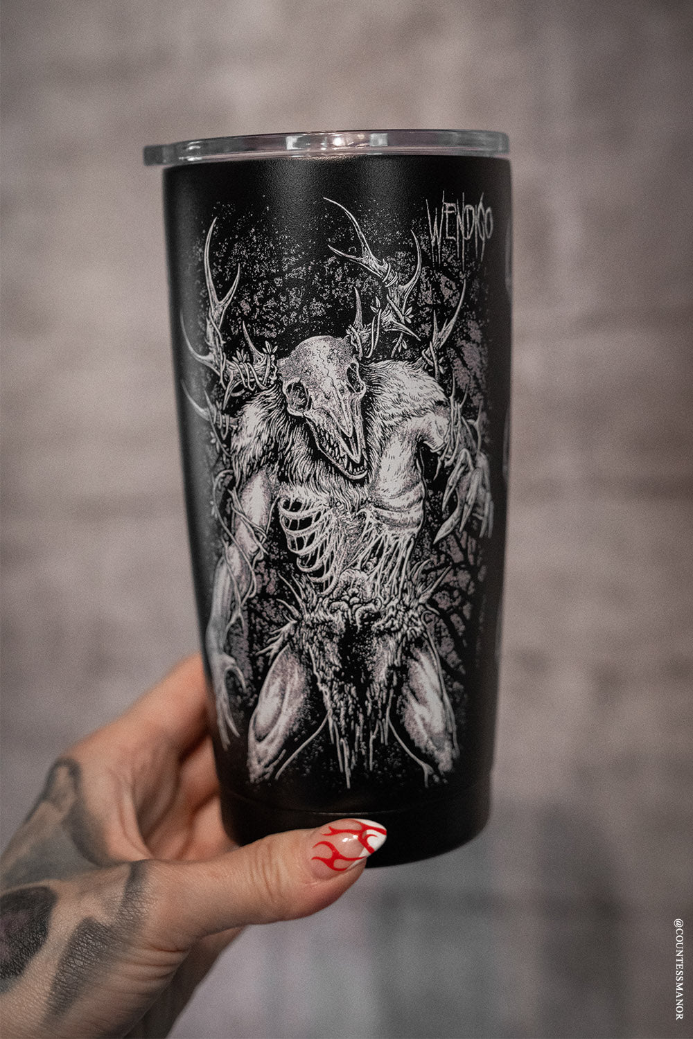 Black tumbler with a detailed skull and deer design held by a hand with red nail polish.