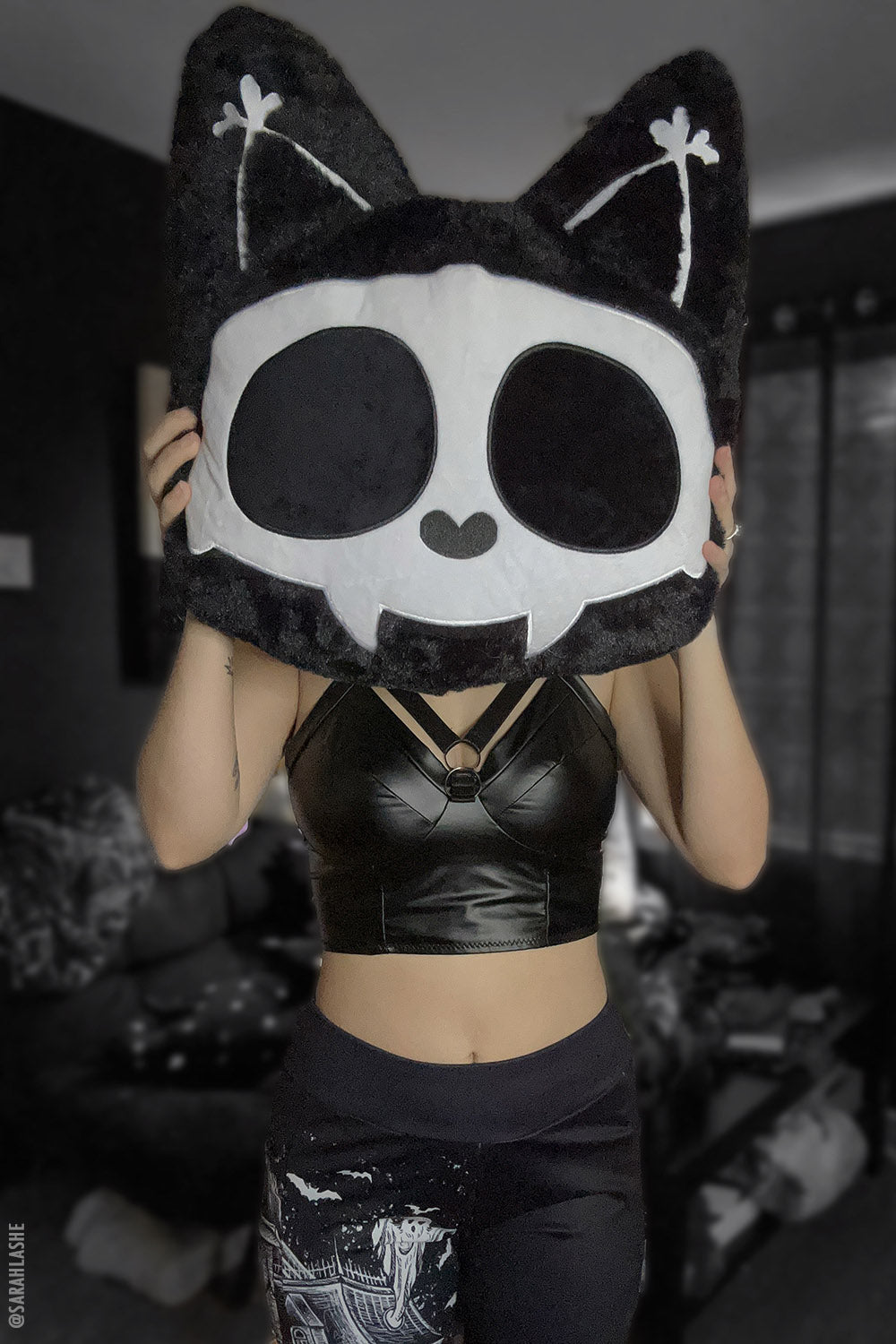Person holding a large throw pillow with a 3D cat shape featuring white skeleton bones on a black background with embroidered details.