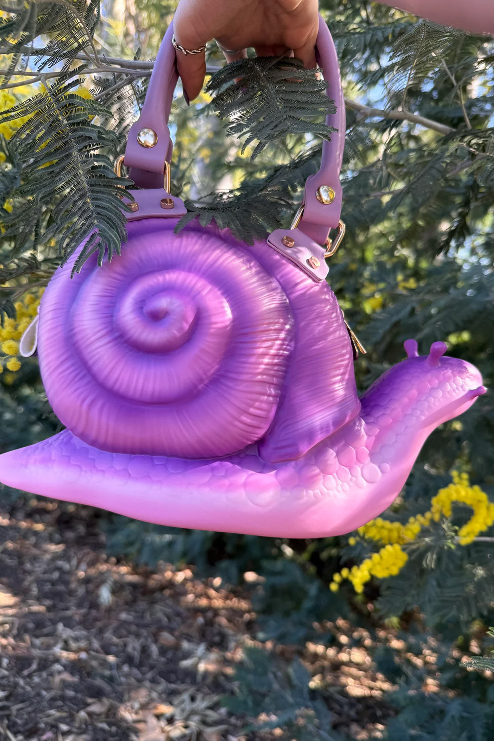 Purple snail-shaped handbag held by a hand with greenery and flowers in the background