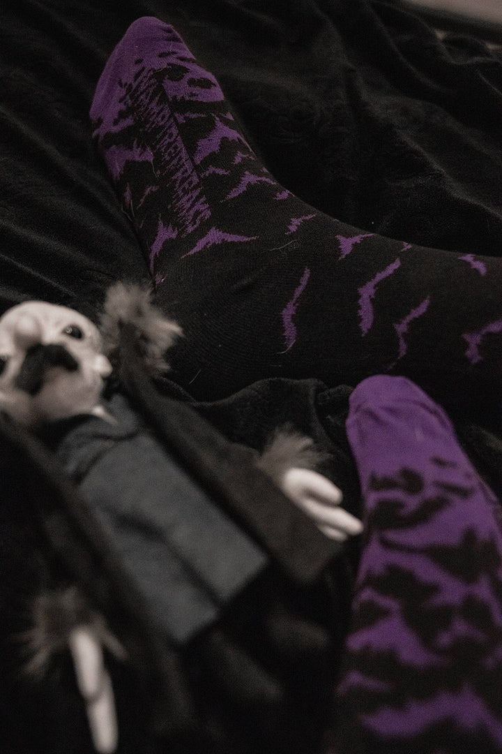Person wearing purple patterned bat socks sitting on a dark surface with a small vampire doll