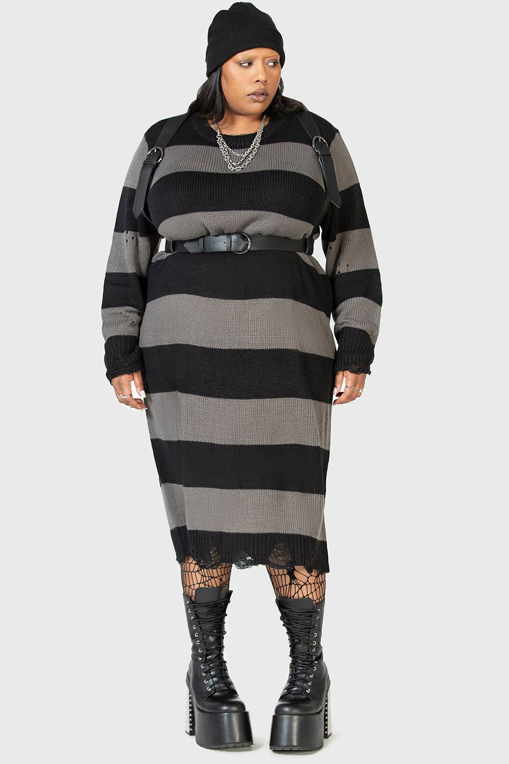goth girl wearing a black and gray striped emo dress with a high-waisted plus size gothic belted harness and platform boots on a white background