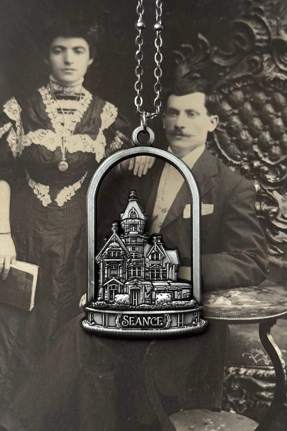 Vintage-style gothic haunted house necklace pendant with haunted house design held by a man and woman in formal attire.