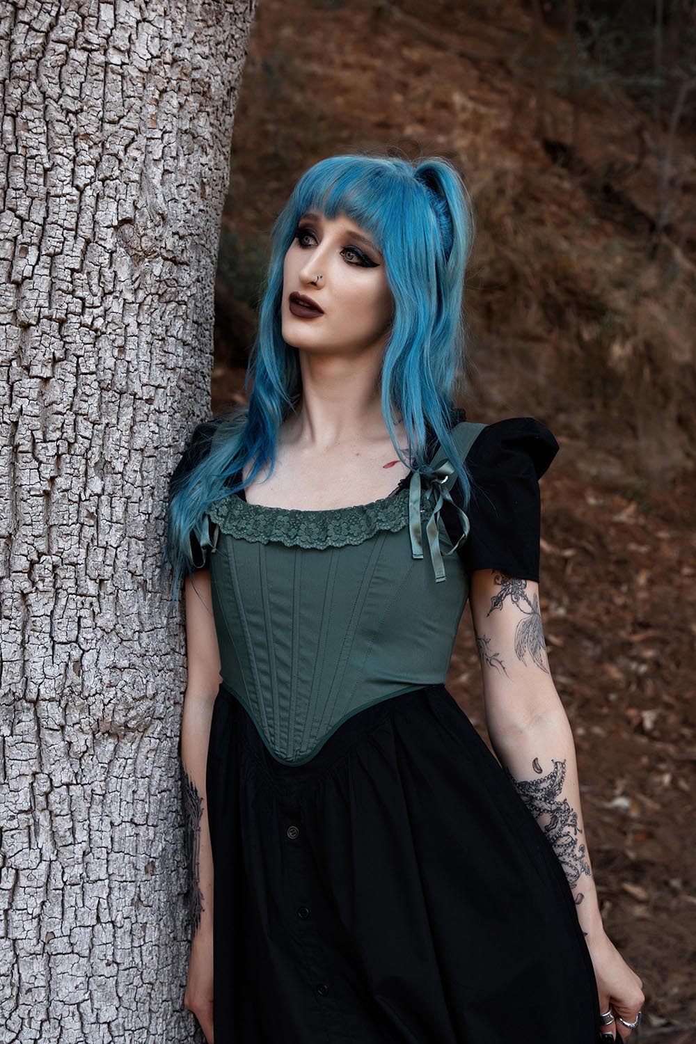 Person with blue hair leaning against a tree in a forest setting wearing a corset top