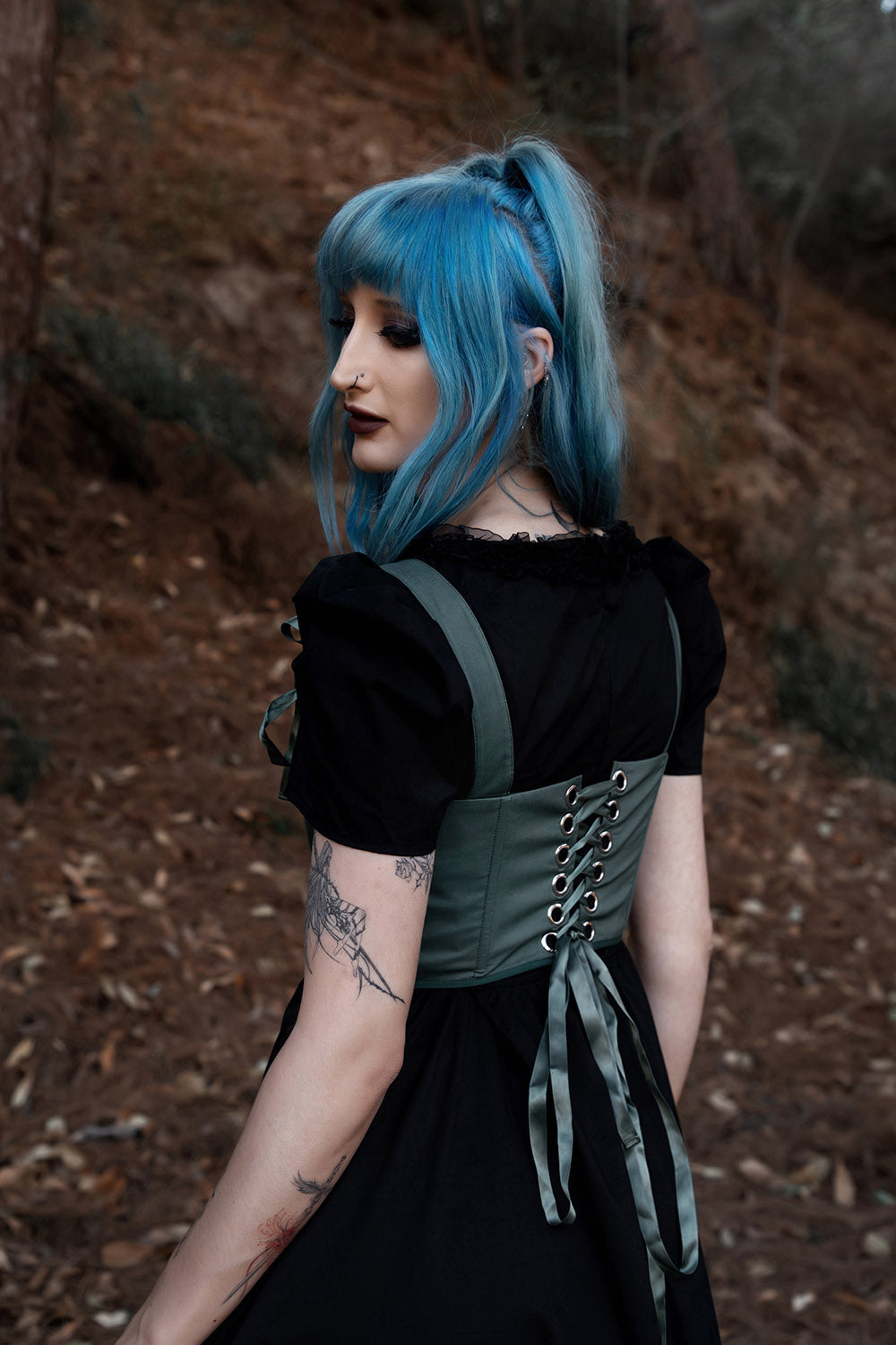 Person with blue hair wearing a black dress with green corset in a forest setting